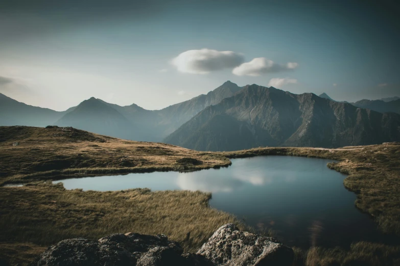 a body of water sitting on top of a grass covered hillside, by Sebastian Spreng, pexels contest winner, tall mountains in the horizon, screensaver, mini lake, with mountains in the background