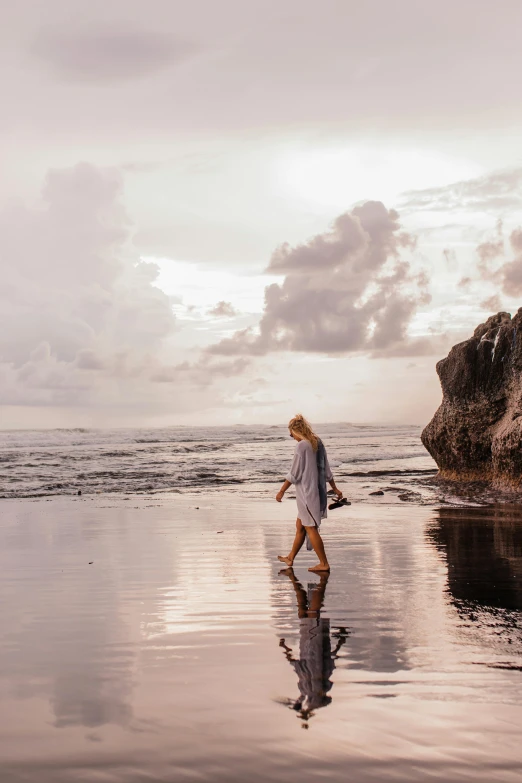 a woman walking on a beach next to a rock, unsplash contest winner, bali, glossy reflections, late summer evening, grey