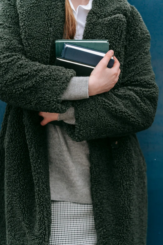 a woman standing in front of a blue wall holding a book, by James Morris, trending on unsplash, wearing a green sweater, wearing a grey fur robe, piled around, dark green