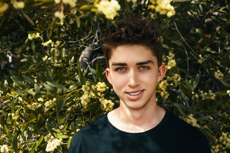 a young man standing in front of a bush of flowers, inspired by John Luke, pexels contest winner, clean shaven wide face, kailee mandel, around 20 yo, avatar image