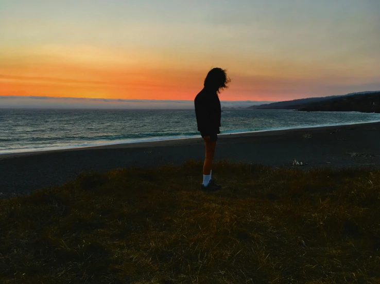 a woman standing on top of a beach next to the ocean, an album cover, unsplash contest winner, orange lit sky, standing alone in grassy field, hollister ranch, profile image
