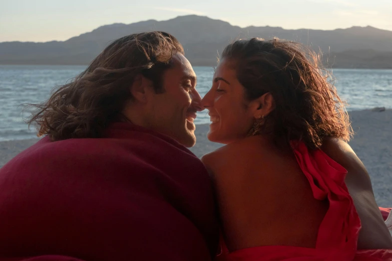 a man and a woman sitting next to each other on a beach, pexels contest winner, happening, red cloth around his shoulders, justina blakeney, profile image, covered with blanket