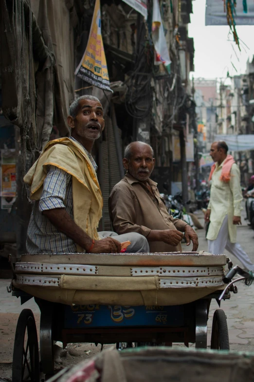 a couple of men riding on the back of a cart, inspired by Steve McCurry, trending on unsplash, dressed in a jodhpuri suit, bustling city, sitting down, grey