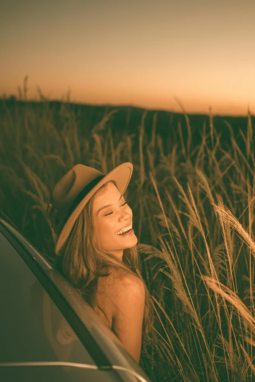 a woman sitting in the grass next to a car, trending on pexels, sunset glow around head, huge smile, with hat, fields