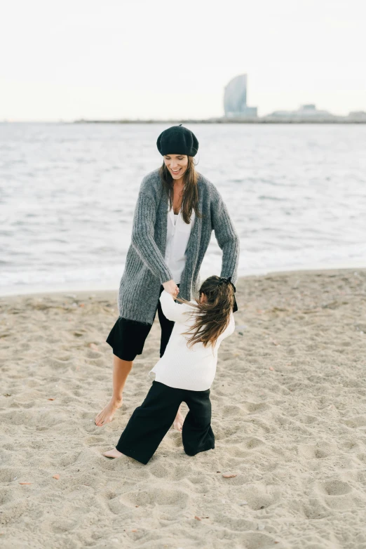 a woman standing next to a little girl on a beach, wearing in cardigan, in barcelona, grey, playful pose