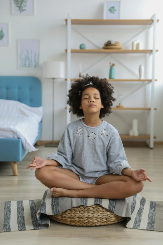 a woman sitting in the middle of a room doing yoga, a portrait, by Everett Warner, pexels, light skinned african young girl, inside a child's bedroom, meditating pose, wearing a grey robe