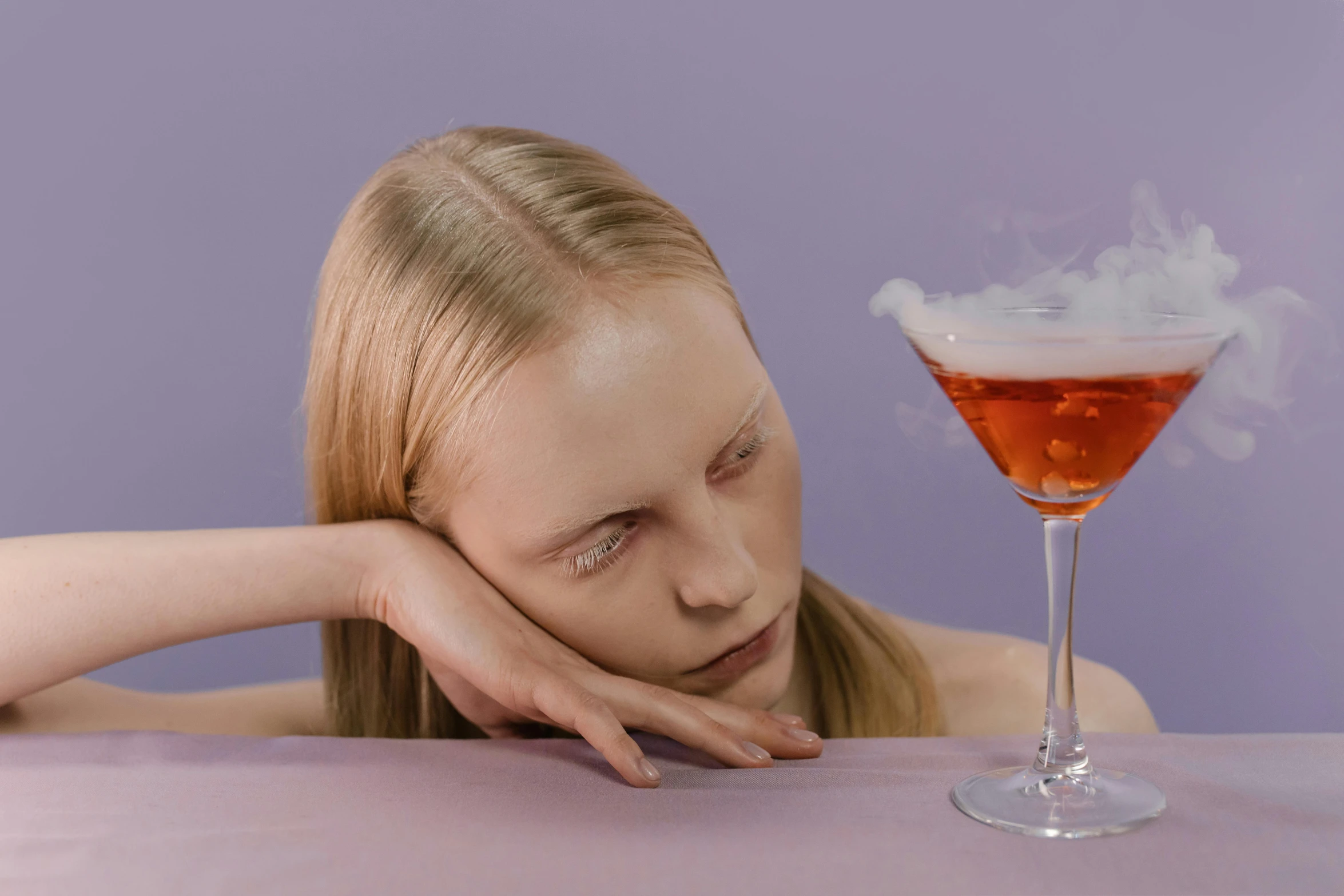 a woman sitting at a table with a drink in front of her, an album cover, inspired by Méret Oppenheim, trending on unsplash, magic realism, dry ice, sleepy fashion model face, on a pale background, people drink cocktails