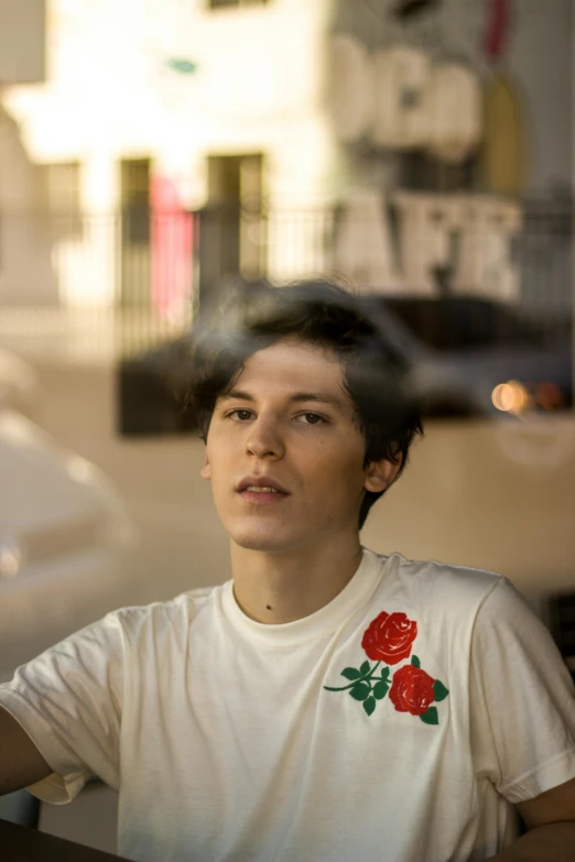 a man sitting at a table in front of a window, an album cover, by Elsa Bleda, trending on unsplash, portrait androgynous girl, andy samberg, handsome face, photo of a rose