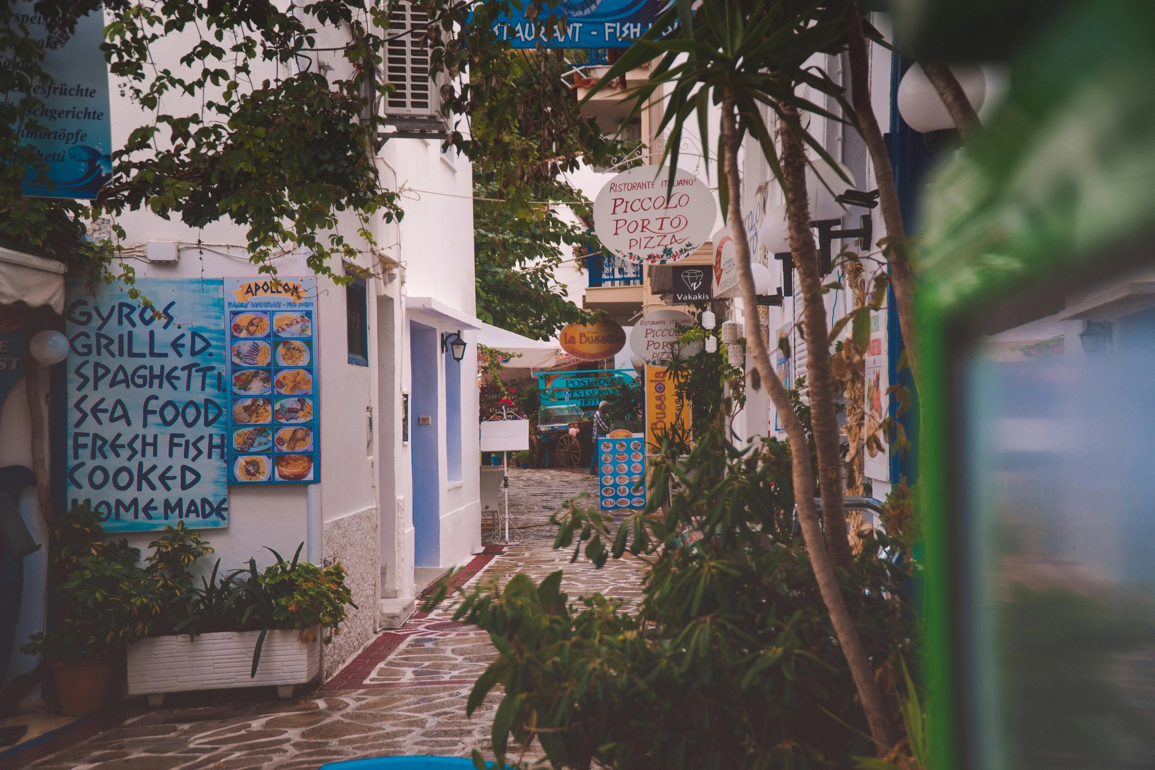 a narrow street with a bunch of signs on it, by Julia Pishtar, greek nose, background image, tropical coastal city, thumbnail