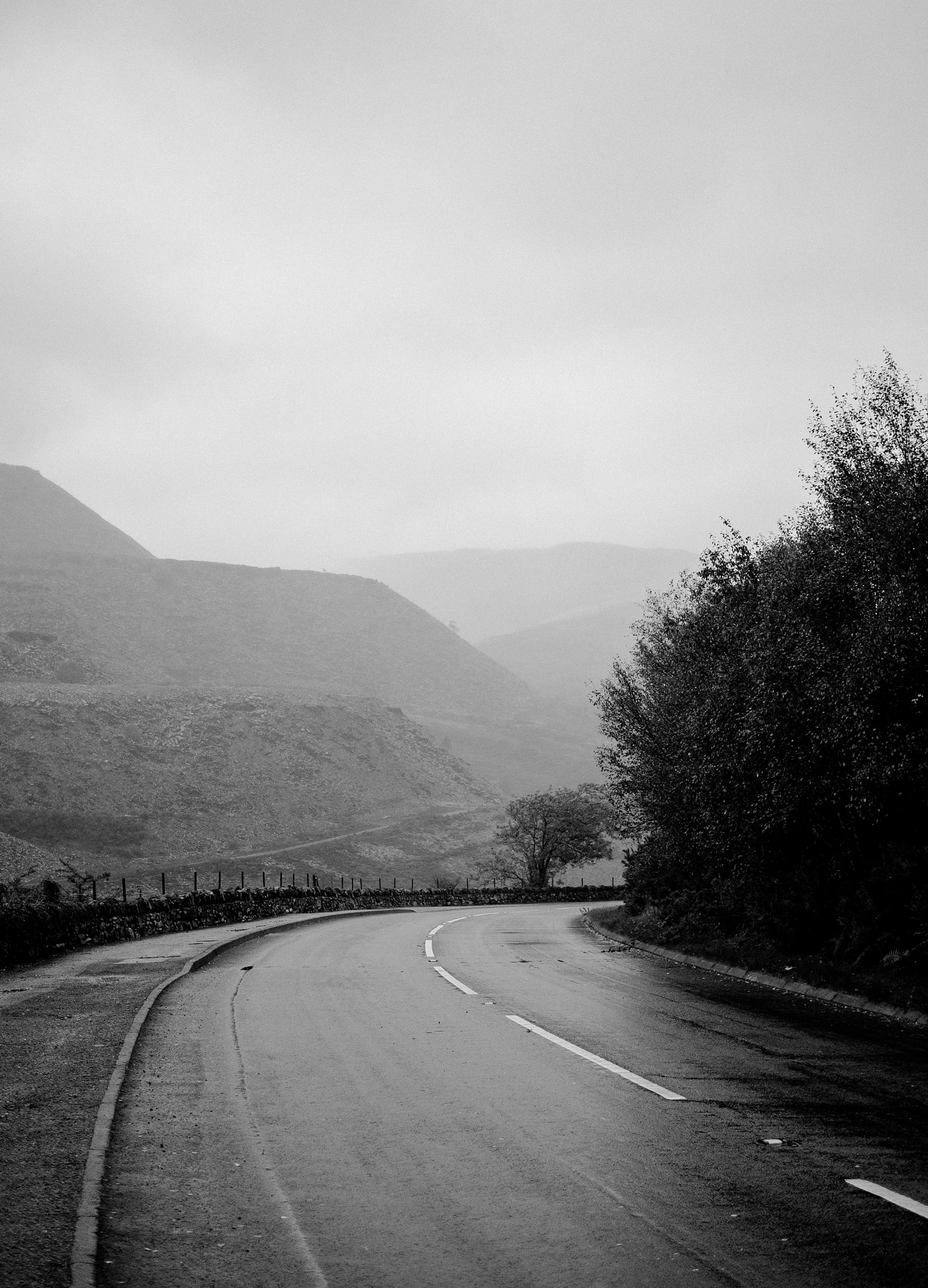 a black and white photo of an empty road, by Andrew Robertson, unsplash, hills in the background, rain and haze, marsden, autumnal