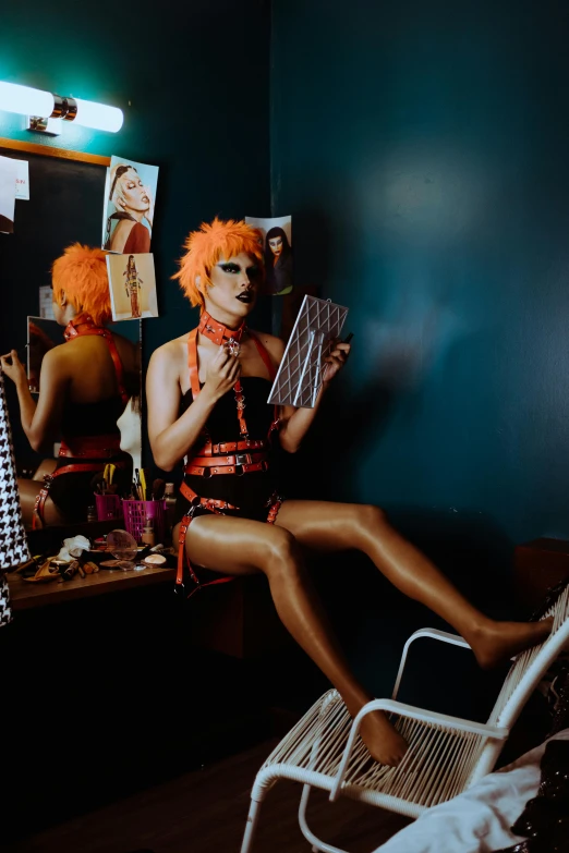 a woman sitting in a chair in front of a mirror, an album cover, inspired by Nan Goldin, trending on pexels, conceptual art, dressed in orange inmate attire, clown makeup and rainbow wig, theater dressing room, drag