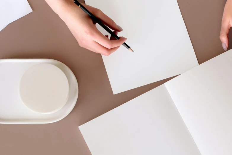 a person holding a pen and writing on a piece of paper, by Julia Pishtar, trending on pexels, white ceramic shapes, sitting on a mocha-colored table, full view blank background, white panels