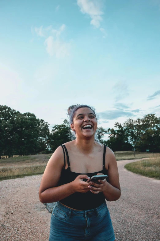 a woman standing on a dirt road holding a cell phone, both laughing, she is wearing a black tank top, 2019 trending photo, sydney park