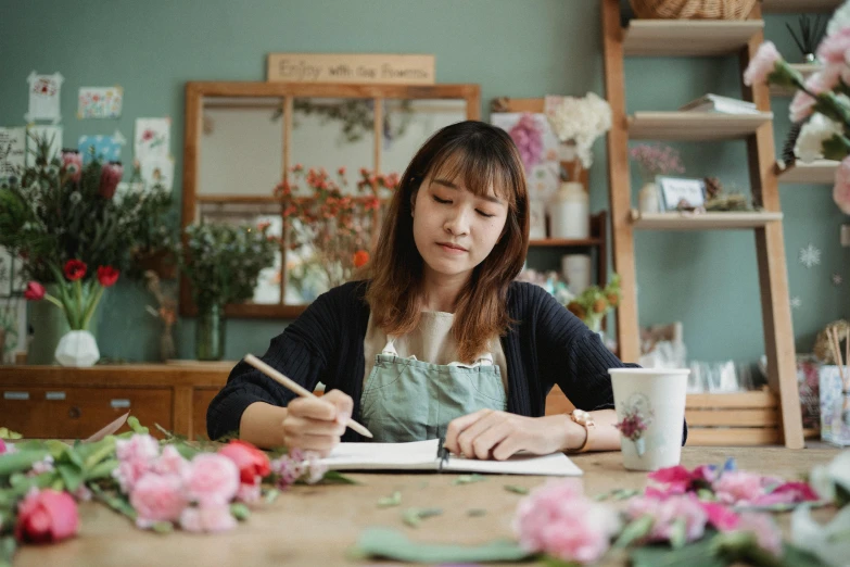 a woman sitting at a table writing on a piece of paper, inspired by Ruth Jên, pexels contest winner, flower shop scene, avatar image, maintenance, cutest