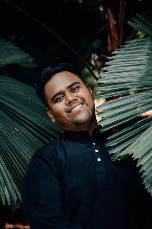 a man standing in front of a palm tree, an album cover, pexels contest winner, hurufiyya, headshot portrait, south east asian with round face, wearing a black shirt, post graduate