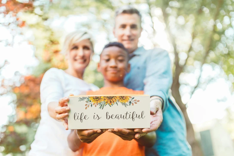 a man and two women holding a sign that says life is beautiful, by Will Ellis, pexels contest winner, holding gift, husband wife and son, rectangle, handcrafted