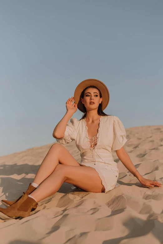 a woman sitting on top of a sandy beach, by Robbie Trevino, stylish pose, at the desert, with hat, gemma chen