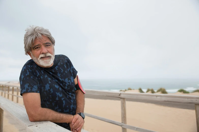 a man standing on a boardwalk next to the ocean, a portrait, by Alejandro Obregón, some grey hair in beard, dunes in the background, george lopez, 15081959 21121991 01012000 4k