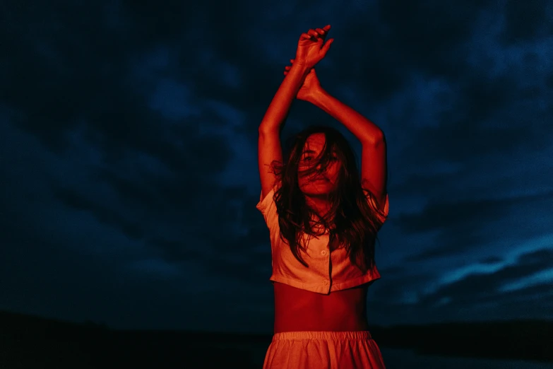 a woman standing in the middle of a field at night, an album cover, by Emma Andijewska, unsplash contest winner, red tank top and wide blue pants, captures emotion and movement, red glowing skin, hands in her hair