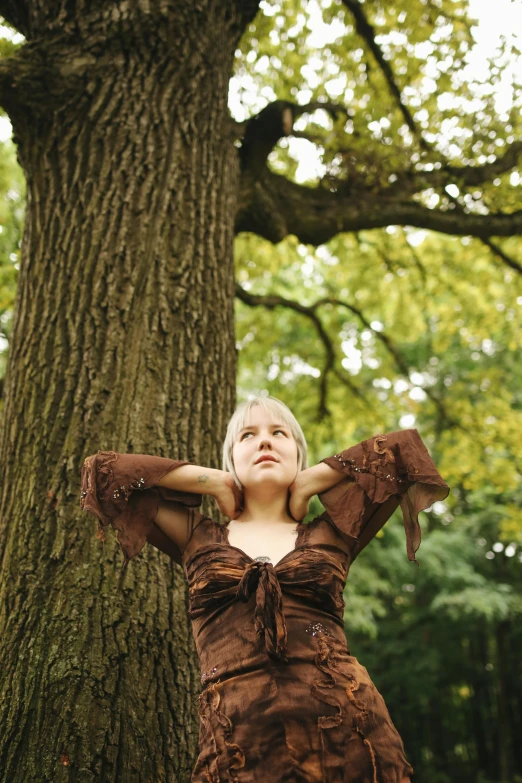 a woman in a brown dress standing next to a tree, inspired by Jane Nasmyth, unsplash, renaissance, a blond, shrugging arms, medium format, concert