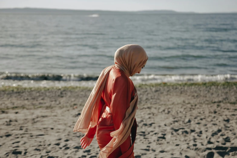 a woman walking on a beach next to the ocean, unsplash, hurufiyya, hijab, flowing salmon-colored silk, seattle, still from the movie