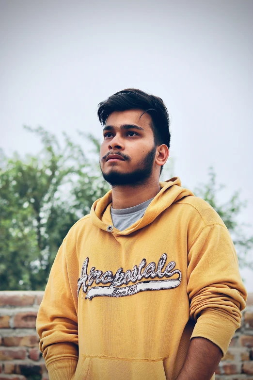 a man standing in front of a brick wall, inspired by Saurabh Jethani, pexels contest winner, wearing a yellow hoodie, profile image, at college, loveable guy