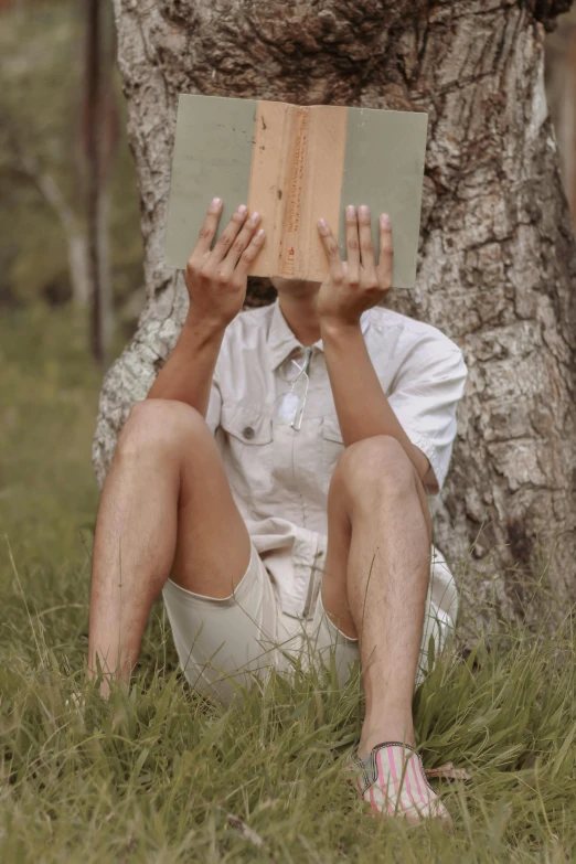 a man sitting under a tree reading a book, tan skin a tee shirt and shorts, holding his hands up to his face, wearing a linen shirt, holding a wood piece