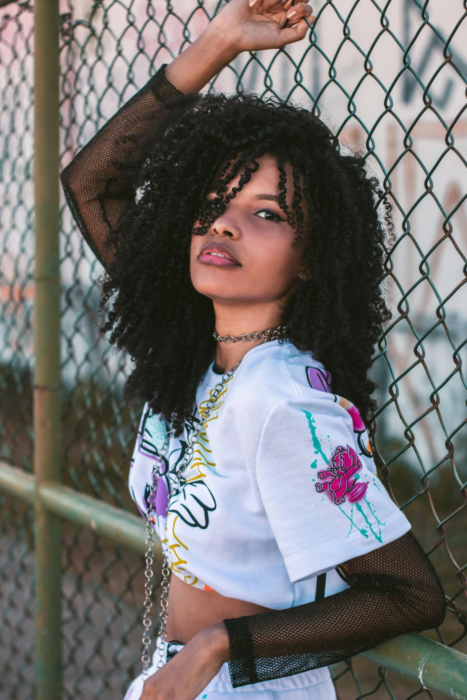 a woman leaning against a chain link fence, a portrait, trending on pexels, graffiti, (dark shorter curly hair), graphic tees, flowery, african american young woman