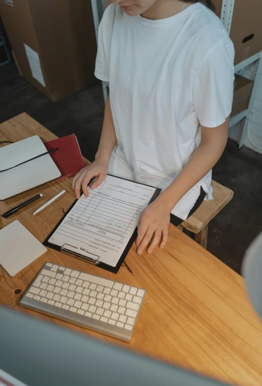 a person sitting at a table with a laptop and a keyboard, holding a clipboard, wearing white shirt, wooden desks with books, high-quality photo