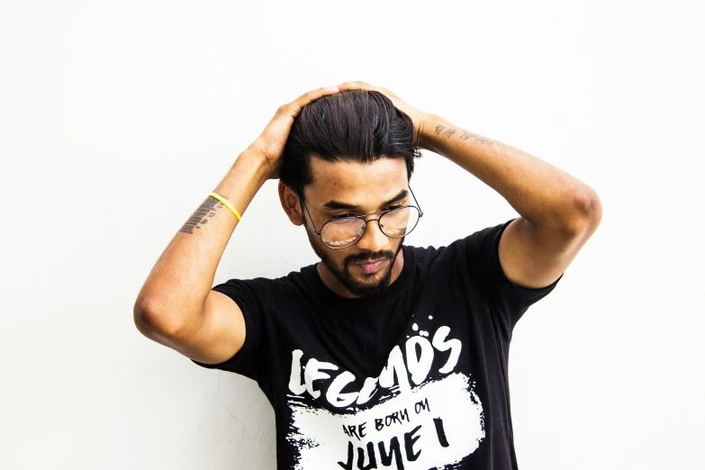 a man standing in front of a white wall with his hands on his head, inspired by Saurabh Jethani, trending on pexels, black t shirt, messy manbun, with glasses on, he is a long boi ”