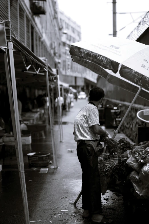 a black and white photo of a man standing under an umbrella, by Gang Hui-an, unsplash, getting groceries, some stalls, taken in the late 2000s, wet market street