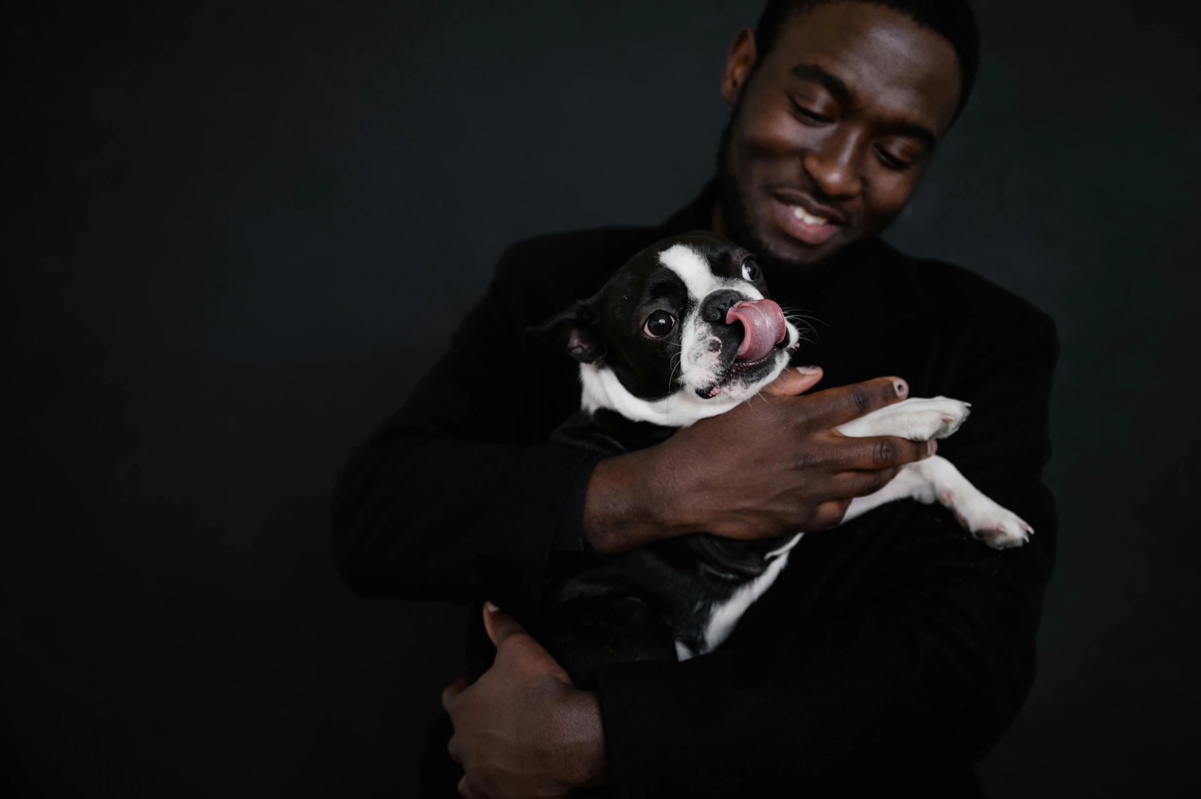 a man holding a black and white dog in his arms, by Emma Andijewska, pexels contest winner, man is with black skin, plain background, jaylen brown, friendly guy and small creature