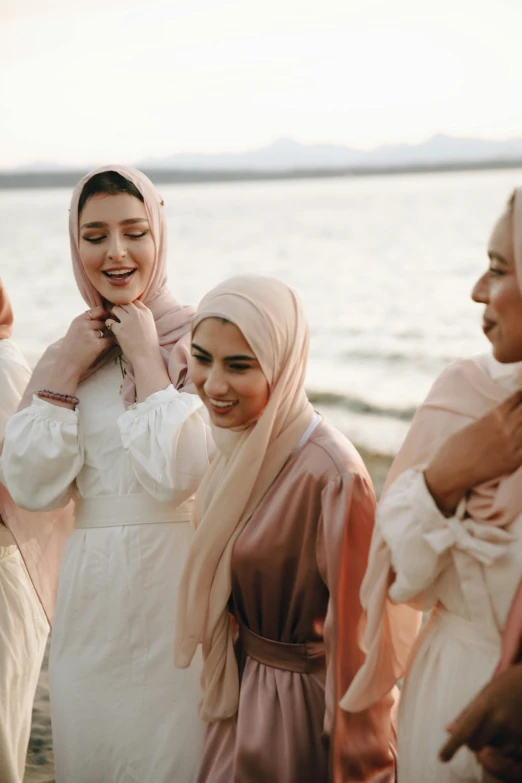 a group of women standing next to each other on a beach, inspired by Maryam Hashemi, trending on unsplash, hurufiyya, white hijab, turning her head and smiling, silk robes, on a lake