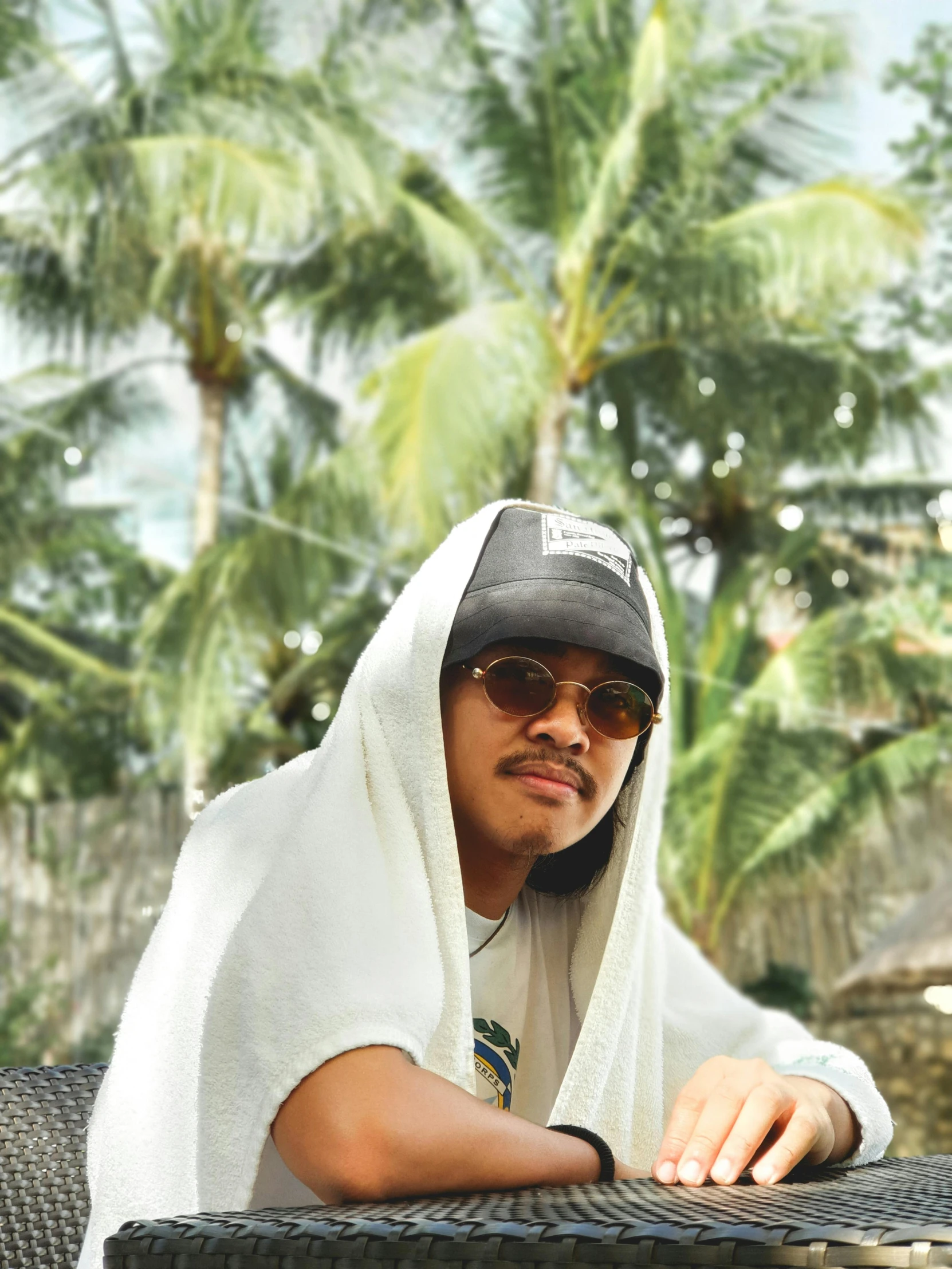 a man sitting on top of a wicker chair, an album cover, inspired by Byron Galvez, unsplash, realism, wearing a white bathing cap, with palm trees in the back, unibrow, ☁🌪🌙👩🏾