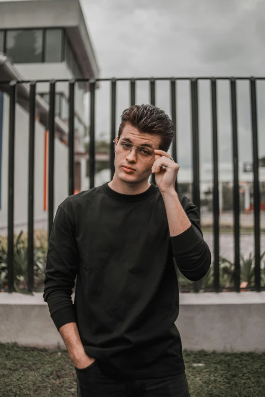 a man standing in front of a fence talking on a cell phone, inspired by John Luke, trending on pexels, realism, wearing a black sweater, wearing black glasses, posing for a picture, brown haired