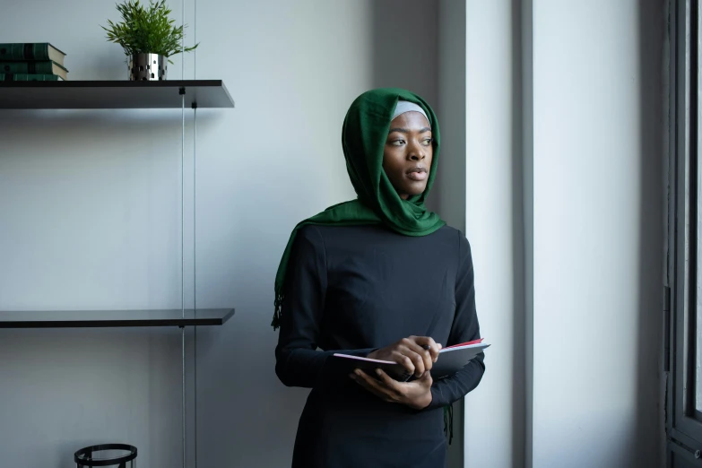 a woman standing in front of a window holding a book, pexels contest winner, hurufiyya, black and green scheme, working in an office, muslim, looking serious