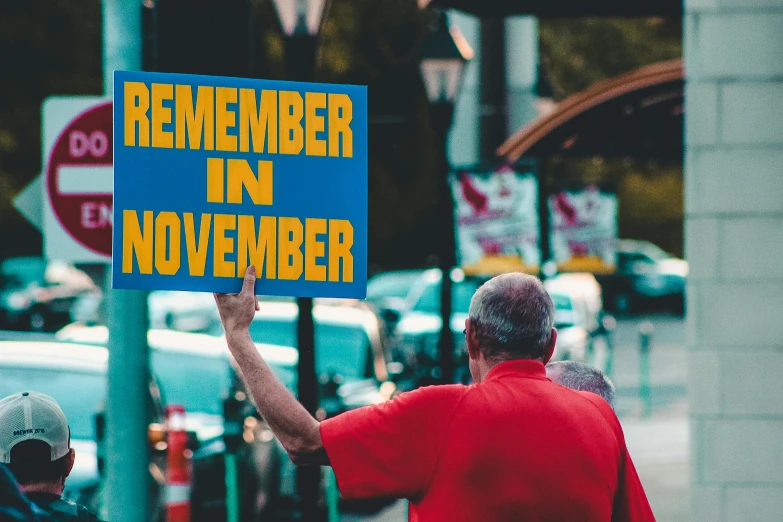 a man holding a sign that says remember in november, by Everett Warner, pexels contest winner, happening, joan cornella, remembrance, memphis, thumbnail