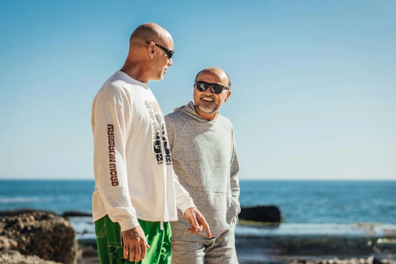 two men standing next to each other on a beach, a portrait, unsplash, portrait of bald, casual streetwear, a green, sun drenched