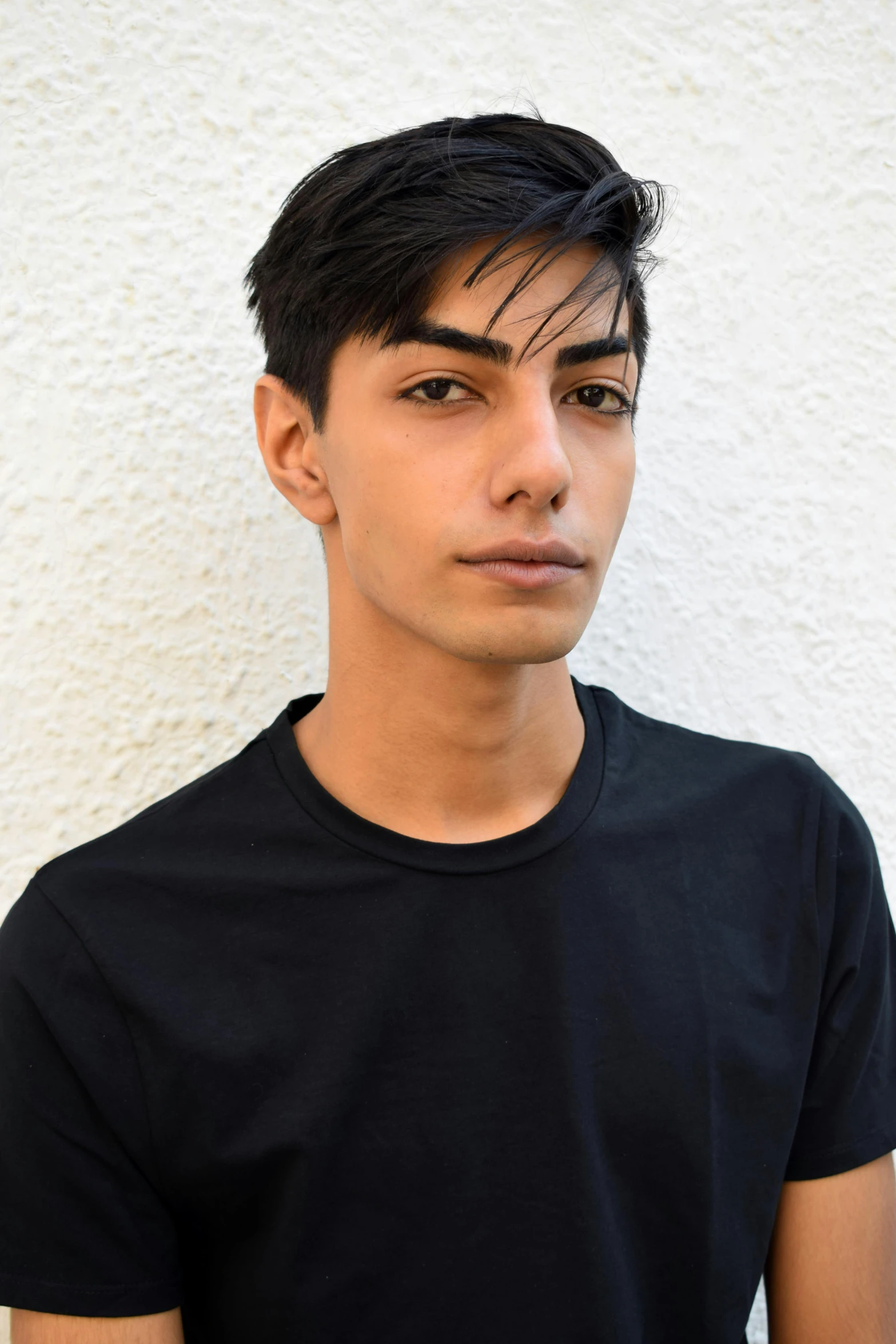 a young man standing in front of a white wall, featured on reddit, androgynous face, middle eastern, wearing a black shirt, color photo