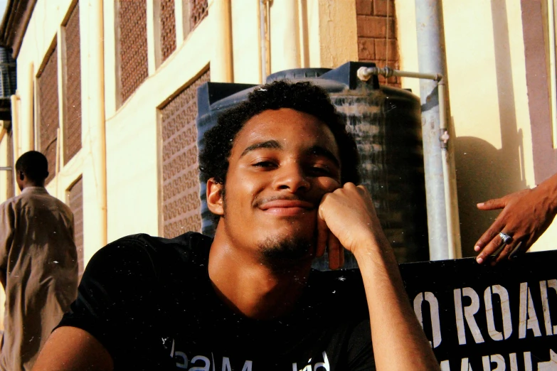 a man sitting in front of a building holding a sign, a picture, by Stokely Webster, pexels contest winner, renaissance, his smile threw shadows, with afro, he is wearing a black t-shirt, profile image