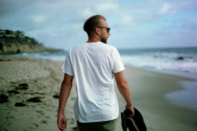 a man standing on a beach holding a skateboard, a polaroid photo, inspired by John E. Berninger, unsplash, minimalism, blond brown stubble thin beard, dressed in a white t-shirt, paul giamatti, profile pic