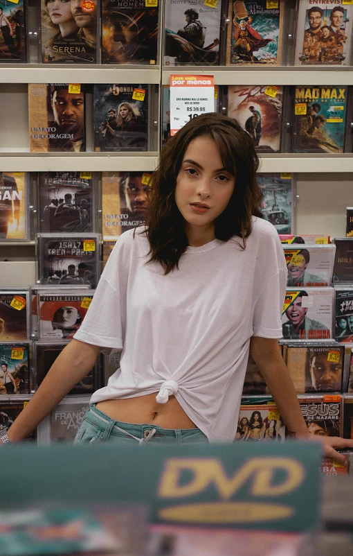 a woman standing in front of a shelf of dvds, inspired by Nan Goldin, pexels contest winner, hyperrealism, dressed in a white t-shirt, chloe bennet, daisy ridley, at a mall