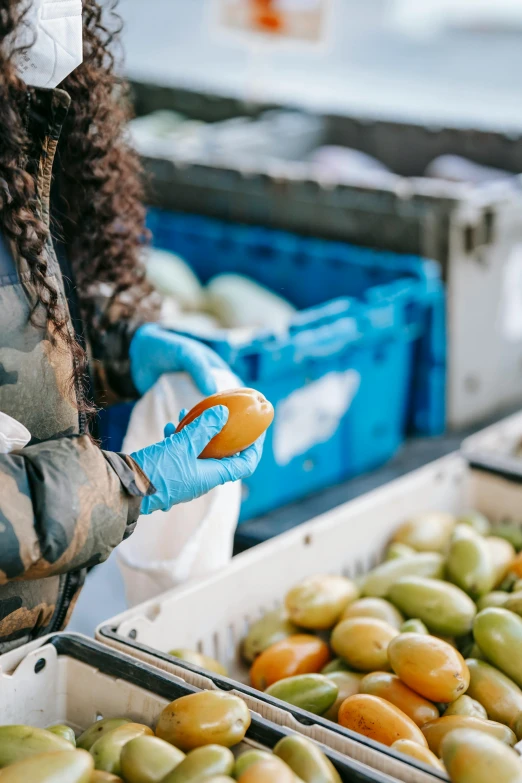 a woman wearing a face mask standing next to boxes of fruit, arbeitsrat für kunst, blue gloves, north island brown kiwi, busy market, thumbnail