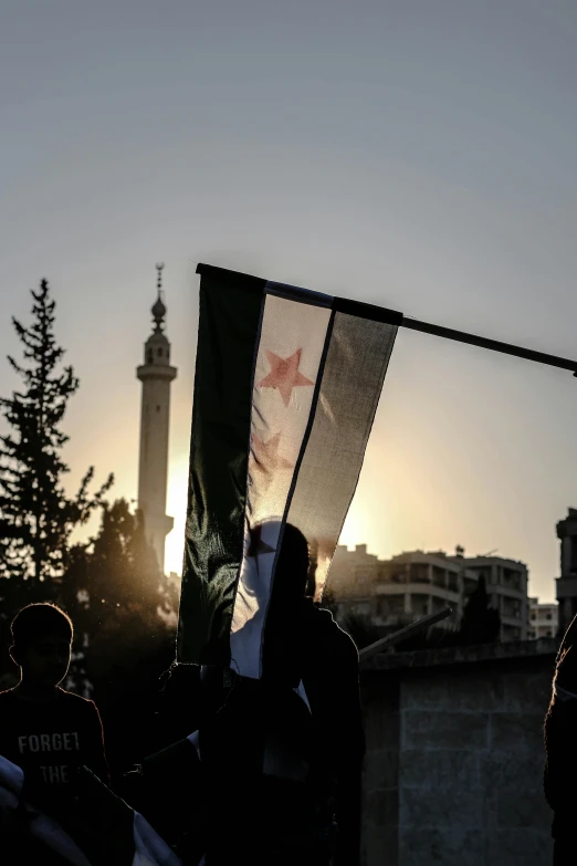 a group of people standing in front of a flag, by Cafer Bater, trending on unsplash, hurufiyya, real life photo of a syrian man, evening sun, ( ( theatrical ) ), spire