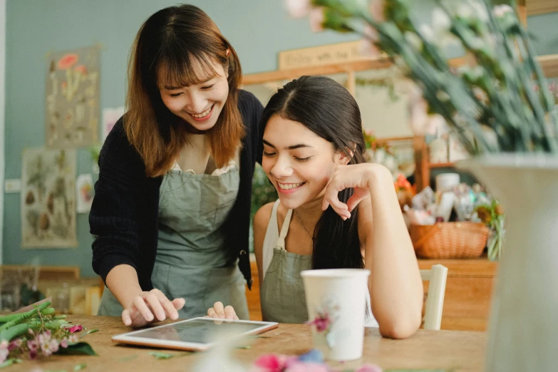 two women in a flower shop looking at a tablet, a cartoon, trending on pexels, sitting on a mocha-colored table, avatar image, looking her shoulder, student