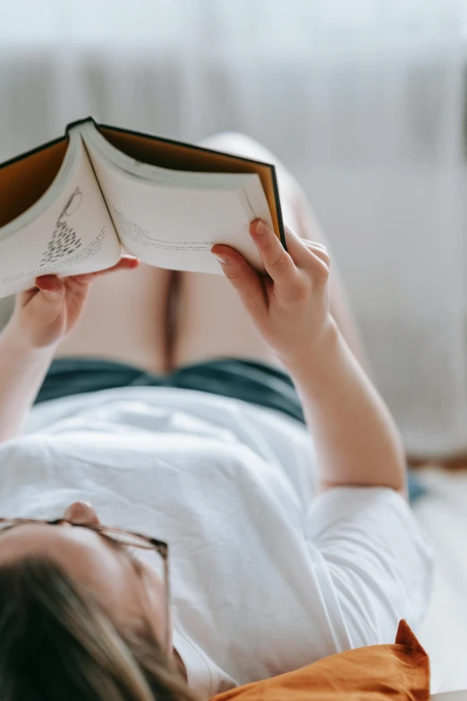 a woman laying on a bed reading a book, wearing a white button up shirt, large thighs, short bookshelf, zoomed in