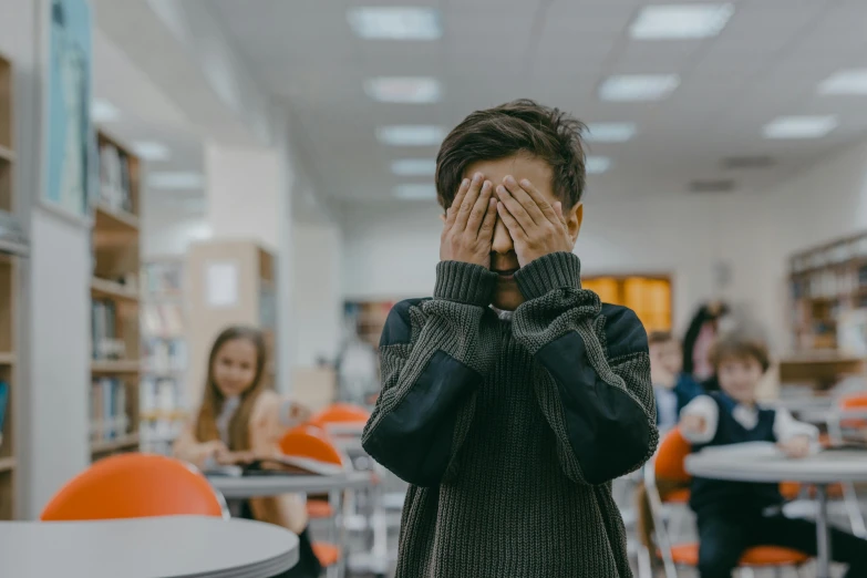 a boy covers his face with his hands in a classroom, pexels contest winner, pixelated, speak no evil, one eye is read, greeting hand on head