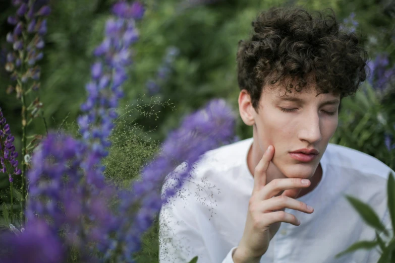 a man sitting in a field of purple flowers, trending on pexels, magic realism, brown curly hair, hand on his cheek, male teenager, androgyny