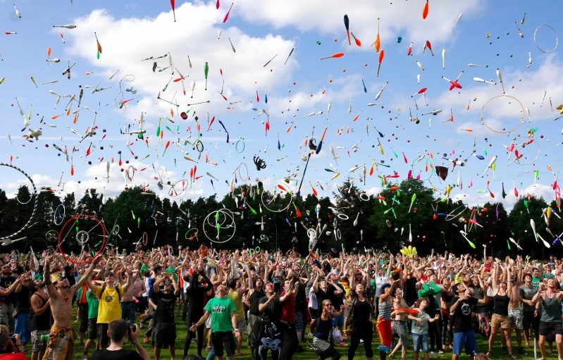 a large group of people standing on top of a lush green field, kites, confetti, hands in the air, revellers