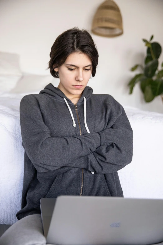 a woman sitting on a bed with a laptop, grey hoodie, frowning, androgynous male, taken in 2 0 2 0